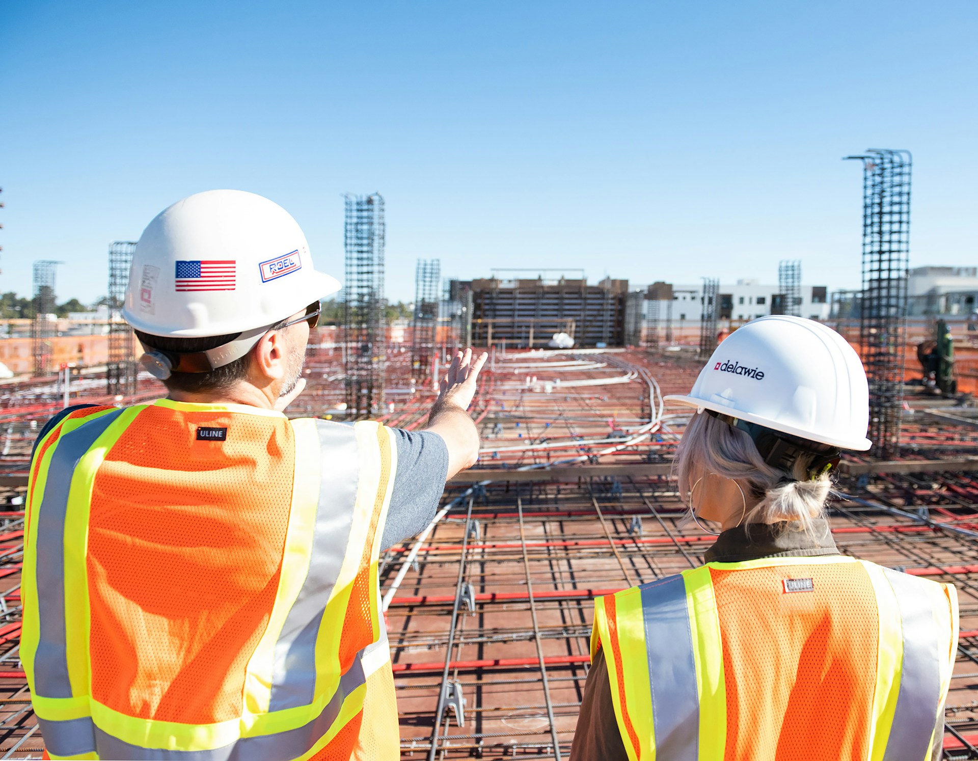 A man and woman wearing hard hats & reflective safety vests while looking over construction site, representing the importance of safety on a construction site.